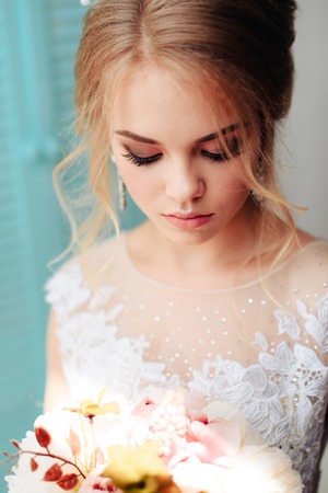 Very beautiful blonde with blue eyes in a white bride dress near a window with a bouquet of flowersの写真素材