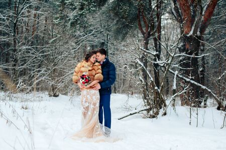Beautiful bride in a fur coat in a winter pine forest with a wedding bouquetの写真素材