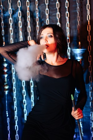 a beautiful brunette girl in a black dress smokes a hookah and lets out smoke 1