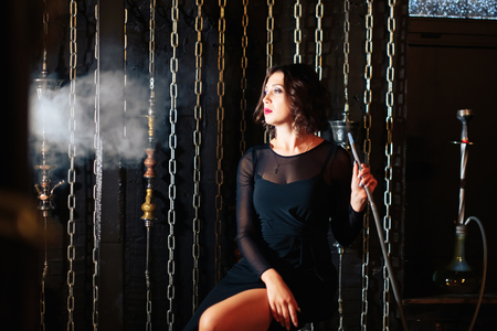 a beautiful brunette girl in a black dress smokes a hookah and lets out smoke 1