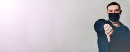 a man in a black medical mask on a gray background gives a thumbs down 1