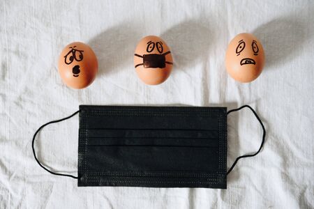 chicken eggs with painted faces on a white cloth background, an egg in a black medical maskの写真素材