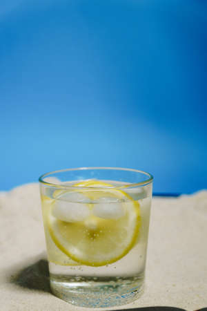 transparent glass with ice and sparkling water on the sand on a blue backgroundの写真素材