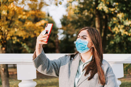 Portrait of a beautiful adult young woman on the background of autumn in the Park in a medical face mask with a phone in her handsの写真素材
