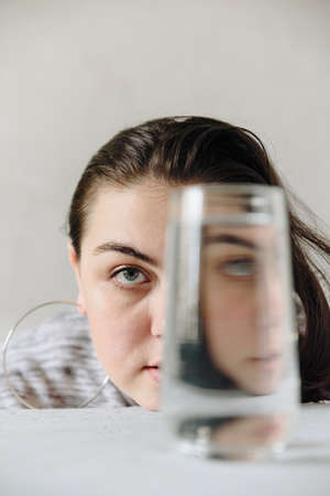 beautiful girl at the table on a gray background in her hands with a glass of water. Girls face through a glass of waterの写真素材