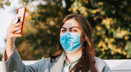 Portrait of a beautiful adult young woman on the background of autumn in the Park in a medical face mask with a phone in her handsの写真素材