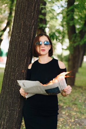 Beautiful woman in a black dress in a park with a burning newspaper in her handsの写真素材