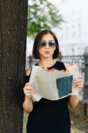 Beautiful woman in a black dress in a park with a burning newspaper in her handsの写真素材