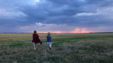 Two children run across a field against the background of clouds at sunset.A boy and a girl in a field against the background of storm cloudsの写真素材
