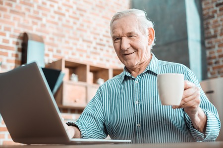 Modern laptop. Cheerful happy senior man smiling and looking at the laptop screen while enjoying new technologyの写真素材