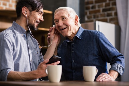Glad to talk to you. Cheerful aged man using smartphone while resting with his son in the kitchenの写真素材