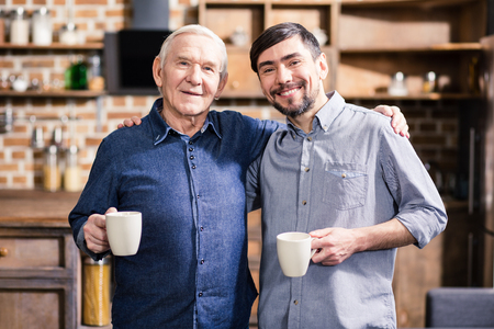 Happy together. Cheerful aged father and his son embracing while drinking coffee at homeの写真素材
