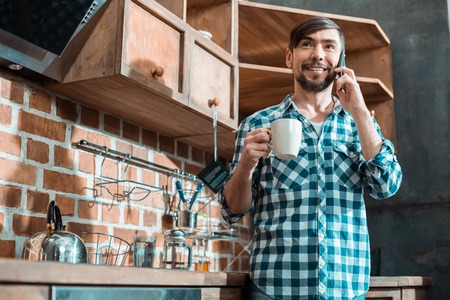 Delicious drink. Cheerful positive nice man smiling and talking on the phone while holding a cup of teaの写真素材