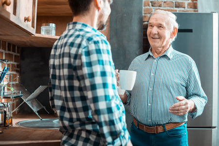 My family. Happy nice elderly man holding a cup of tea and smiling while looking at his sonの写真素材