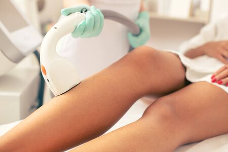 Close up photo of the hand holding modern laser hair removal and putting it on the leg of womanの写真素材