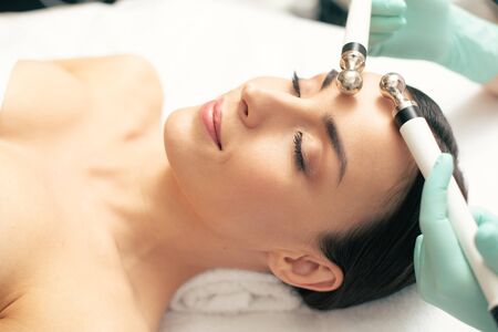 Dark haired lady smiling with her eyes closed and feeling relaxed while having microcurrent therapy in the clinicの写真素材