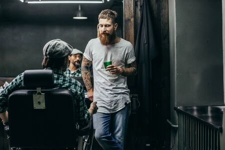 Thoughtful bearded barber looking at his client and holding glass