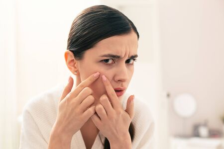 Serious woman squeezing pimple on her face and frowningの写真素材