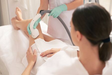 Woman in white bathrobe sitting on the medical couch and undergoing laser hair removal with a vacuum...