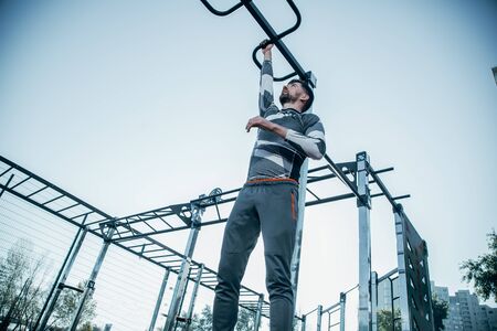 Serious strong sportsman looking up while putting one hand on the chin up bar while hanging from it