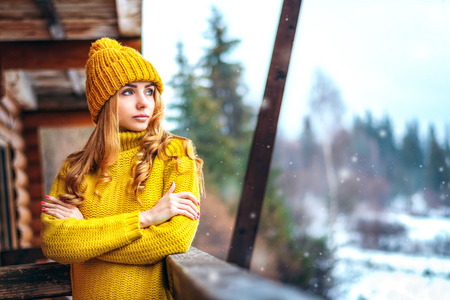 Blond girl in a yellow knit sweater and cap, folded her arms and looking at the snow. Country house in the forest, wooden veranda. cold winter weather. Place for textの写真素材