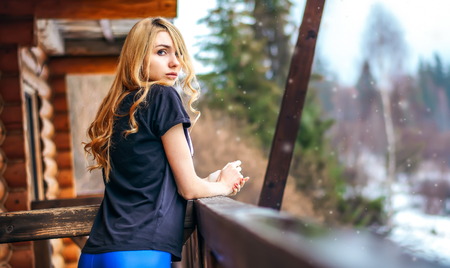 beautiful girl standing on the porch of a wooden house in the winter forest and looks. Long blonde curly hair, snowingの写真素材