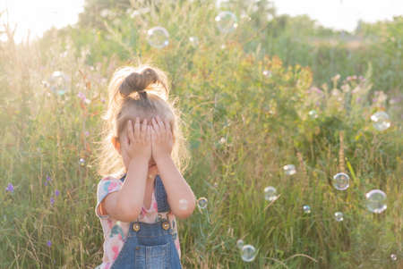 little girl covers her eyes with her hands and stands among the soap bubblesの写真素材