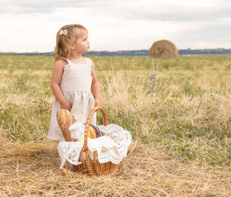 toddler girl in linen dress in field of hay with basket of bread and milk.の写真素材