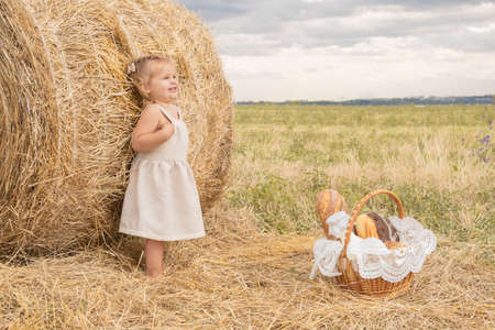 toddler blond girl in linen dress in field of hay with basket of bread and milk.の写真素材
