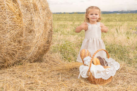 toddler girl blond with basket of bread stands in a field with a haystack.の写真素材