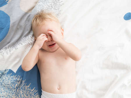 Cute toddler boy in diapers lies on bed rubs his eyes.