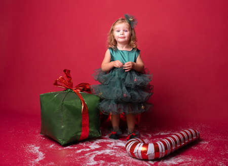 Toddler girl in green Christmas dress with Xmas candy cane and gifts on a red background.の写真素材