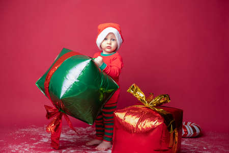 Toddler boy in Santa hat with Xmas gifts and candy cane on a red background.