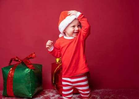 Toddler boy in red Santa hat with Xmas gifts on a red background.