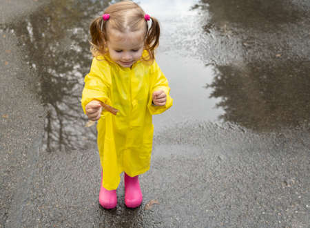 Cute baby girl toddler wearing yellow stylish raincoat pink rubber boots standing in a puddle.の写真素材
