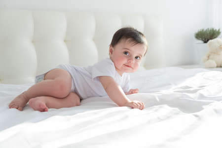 Cute healthy baby girl 6 months smiling in a white bodysuit lying on a bed on white bedding.