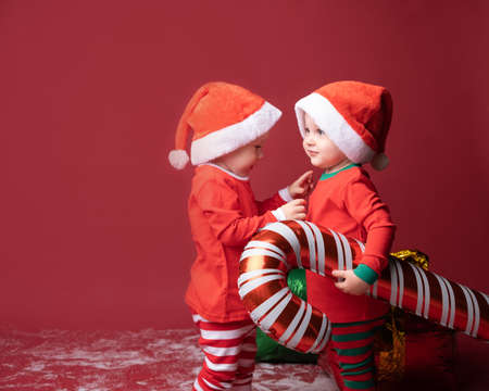 two toddler boys brothers twins in red Santa hat with Xmas candy cane on a red background.
