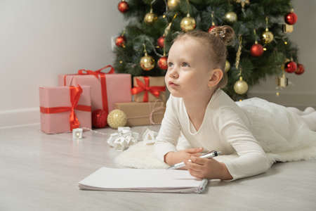 Cute little blonde girl lying on the floor near the Christmas tree writing a letter to Santa Claus.の写真素材