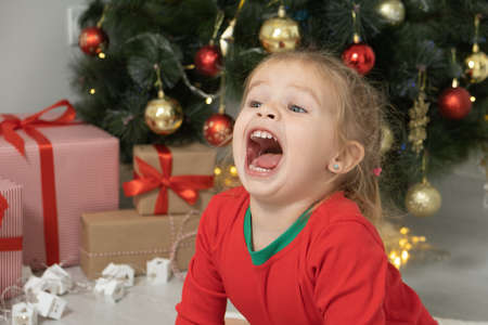 close up portrait excited toddler blonde baby girl in red jacket sitting near the Christmas tree.