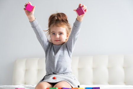Little girl playing with colorful wooden cubes sitting on bed. Home activities for kids.