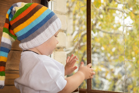 baby boy in a colored hat looks out the window.の写真素材