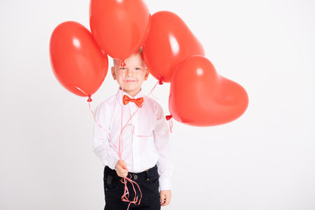 boy in suit and red bow tie holds heart balloons on white backgroundの写真素材