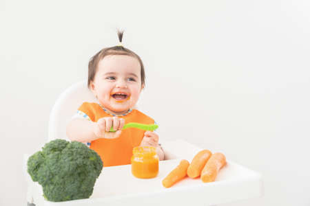 Baby girl in orange bib sitting in a Childs chair eating vegetable puree on white backgroundの写真素材