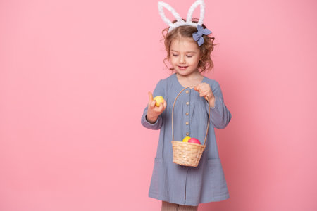 Funny child girl in Easter bunny ears holding basket with colorful eggs on pink background