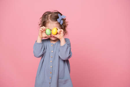 Funny cute child girl in blu dress holding up a colorful Easter egg in front of her eye on pink backgroundの写真素材
