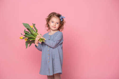 beautiful little blonde girl in blu dress with bouquet of yellow tulips on pink background with space for text.の写真素材