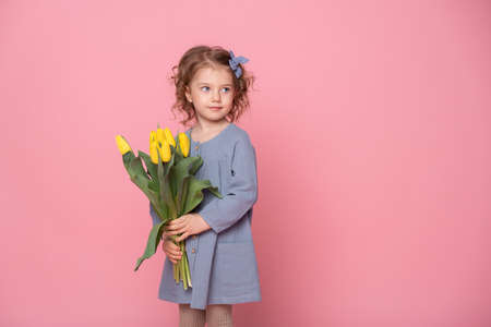 beautiful little blonde girl in blu dress with bouquet of yellow tulips on pink background with space for text.の写真素材
