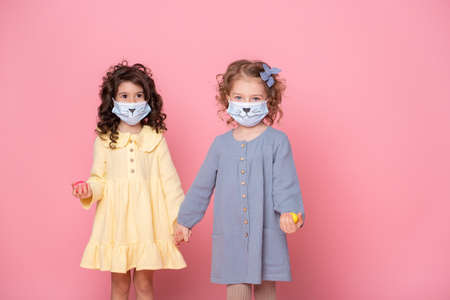 two girls in protective mask with colored eggs holding hands on pink background.の写真素材