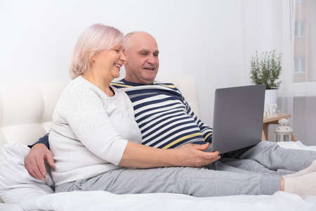 handsome senior couple woman and man sitting in bed using laptop at homeの写真素材