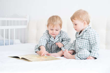 Two toddler baby twin boys in pajamas reading book sitting on white bedding on bedの写真素材